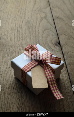 Une boîte cadeau blanche avec couvercle fermé, attaché à un noeud avec ruban vichy rouge et crème. Étiquette vintage vide vers le haut. Banque D'Images