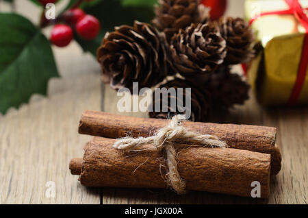 Les bâtons de cannelle, ficelés avec de la ficelle sur une table en chêne avec houx de Noël, des pommes de pins et une boîte cadeau enveloppé d'or dans l'arrière-plan. Banque D'Images