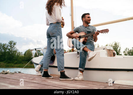 Trois amis se détendre sur pier, assis sur le bateau à voile, man playing guitar Banque D'Images