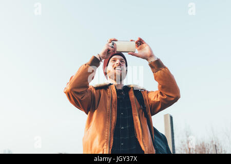 Heureux jeune homme hipster en tenant selfies smartphone contre le ciel bleu Banque D'Images