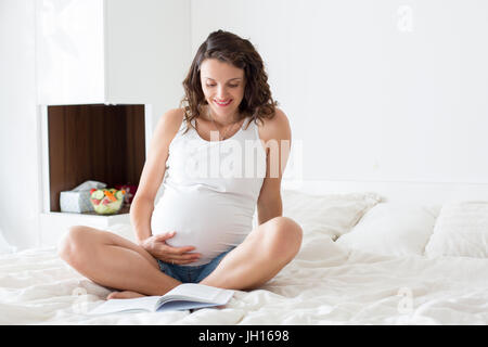 Belle jeune mère enceinte, assis au lit à la maison, manger une salade fraîche et la lecture d'un livre Banque D'Images