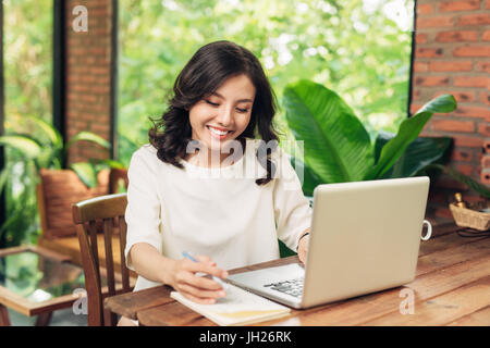 Young business woman sitting at table in cafe et écrit dans l'ordinateur portable. Portable, smartphone et tasse de café sur la table. Banque D'Images