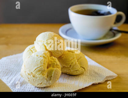 Gros plan sur la bouche-arrosage pain fromage brésilien (Pao de Queijo") sur une serviette en papier blanc, servi avec une tasse de thé noir sur table en bois Banque D'Images