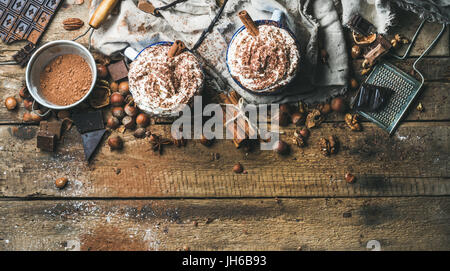 Chocolat chaud avec de la crème fouettée et des bâtons de cannelle servi avec de l'anis étoile, écrous différents et la poudre de cacao sur fond de bois rustique, vue du dessus, art. Banque D'Images