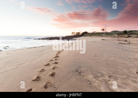 Des traces de pas dans le sable d'une belle plage Australienne mènent à un lever de soleil rose coloré. Banque D'Images