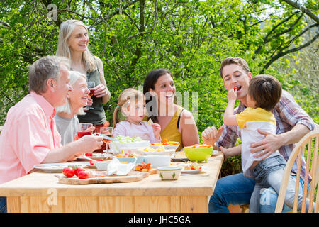 Trois génération de la famille ayant une garden-party à l'été. Tout le monde rit à la recherche et le petit garçon, qui est de nourrir son père certains pe rouge Banque D'Images