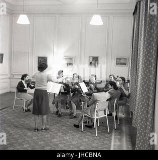 Années 1950, en Angleterre, les étudiants en musique à un collège de formation d'enseignement jouent de leur instrument de musique à l'intérieur d'une chambre dotée de moquette bordée de bord, dirigé par une femme chef d'orchestre. Banque D'Images