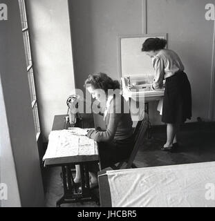 Années 1950, l'Angleterre, deux élèves de sexe féminin à l'intérieur à un collège de formation des enseignants, l'un assis à une petite table à l'aide d'une machine à coudre Singer, conduit l'autre à un lavabo. Banque D'Images