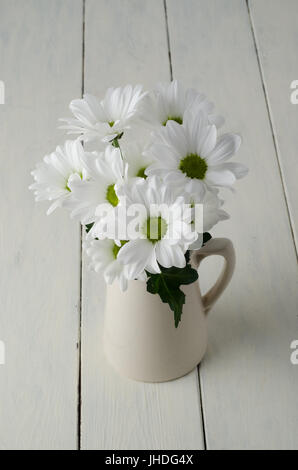 Un petit bouquet de fleurs blanches (chrysanthèmes) avec feuille verte dans un pot à crème beige sur l'extérieur des planches de bois peintes en blanc. Banque D'Images