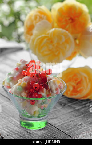 Gâteau blanc avec des fruits confits et de groseille rouge dans une plaque transparente verte sur un fond de roses jaunes.Le cadre vertical. Le cadre vertical. Banque D'Images