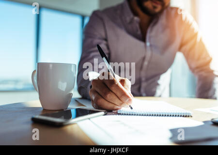 Les investisseurs d'affaires écrit dans le bloc-notes avec une tasse de café sur le bureau. Closeup of a businessman's hand holding pen et prendre des notes. Banque D'Images