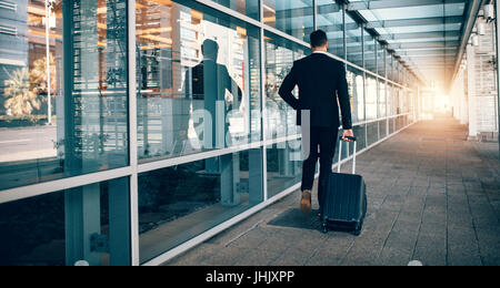 Vue arrière du jeune homme marchant à l'extérieur du bâtiment de transport public avec assurance. Voyageur d'affaires moderne pulling suitcase in airport terminal. Banque D'Images