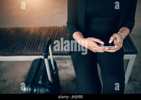 Cropped shot of young businesswoman sitting on bench avec valise et à l'aide de téléphone mobile. Femme en attente dans la station des transports publics. Banque D'Images