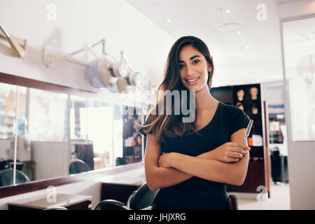 Coiffure femme professionnel à salon avec des ciseaux dans la main. Smiling young woman standing in salon avec mains jointes. Banque D'Images