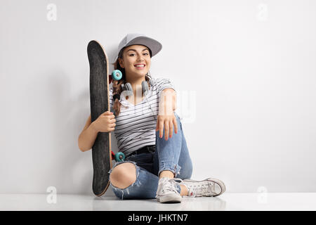 Teenage girl with a skateboard et écouteurs assis sur le plancher et appuyé contre un mur Banque D'Images