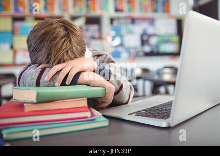 Écolier fatigué se reposant sur la pile de livre par laptop in classroom Banque D'Images