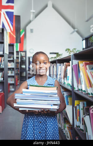 Portrait of smiling girl smiling tout en portant des livres par étagère de bibliothèque Banque D'Images