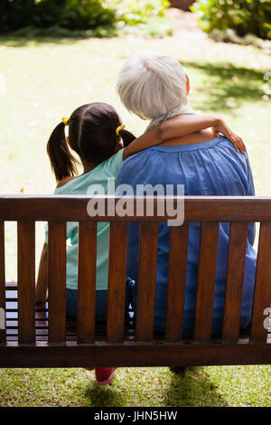 Vue arrière de fille avec bras autour de grand-mère assise sur banc en bois à l'arrière-cour Banque D'Images