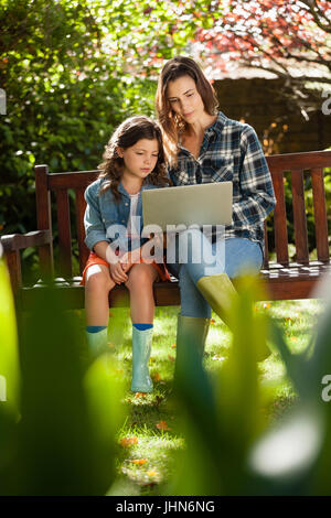 Fille assise par la mère à l'aide de l'ordinateur portable sur banc en bois à l'arrière-cour Banque D'Images