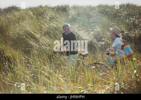 Mature couple walking vélos dans sunny beach grass Banque D'Images