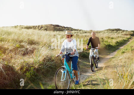 Mature couple riding bicycles on sunny beach chemin d'herbe Banque D'Images