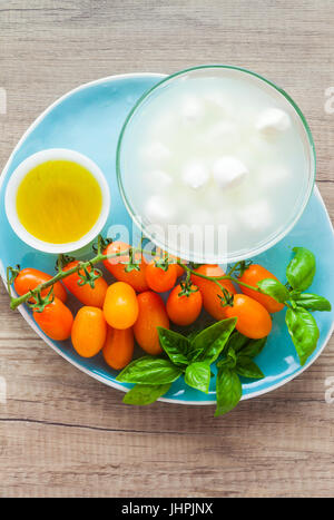 Les petites tomates cerises jaunes et les petits fromages mozzarella à l'huile d'olive et les feuilles de basilic sur une plaque bleue sur une table rustique. Ingrédients pour la salade Banque D'Images