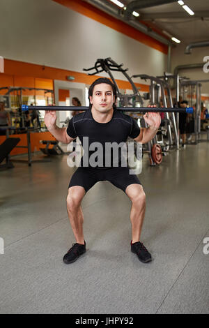 Young man in the black sportwear avec ce barbell squats en salle de sport. Banque D'Images