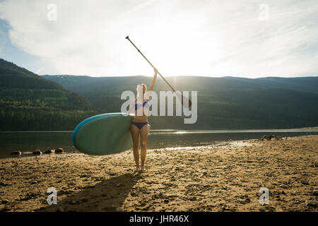 Portrait of smiling young woman standing with paddleboard et oar au lakeshore against sky Banque D'Images