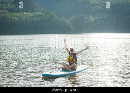 Excitée jeune femme assise avec bras levés sur le lac en paddleboard Banque D'Images