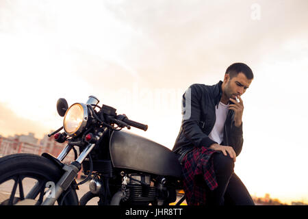 Beau rider homme avec barbe et moustache de biker jacket fumeurs, de penser et de s'asseoir sur un style classique du cafe racer moto au coucher du soleil. Fait sur commande de vélo Banque D'Images