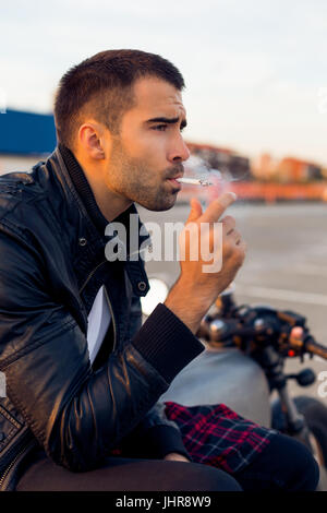 Handsome Guy Rider avec barbe et moustache de biker jacket fumeurs de cigarettes et maintenez-le s'asseoir sur un style classique du cafe racer moto au coucher du soleil. La coutume de vélo m Banque D'Images