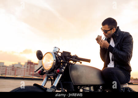 Beau mâle cavalier avec barbe et moustache en noir Veste motard et lunettes de mode 1900 fumée et s'asseoir sur le style classique du cafe racer moto o Banque D'Images