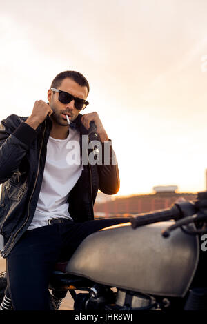 Beau happy rider guy avec barbe et moustache en noir lunettes fashion 1900 fumeurs et corriger biker jacket s'asseoir sur un style classique du Cafe Racer Banque D'Images