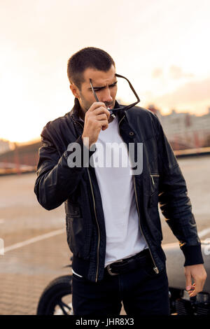 Handsome Guy Rider avec barbe et moustache en noir veste biker, blanc T-shirt et lunettes de 1900 fumeurs près de style classique du cafe racer moto Banque D'Images