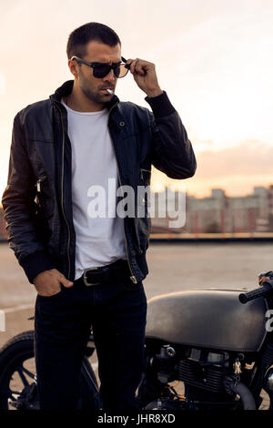 Close up of a handsome guy rider avec barbe et moustache en noir biker jacket and sunglasses 1900 fumeurs près de style classique du cafe racer moto Banque D'Images