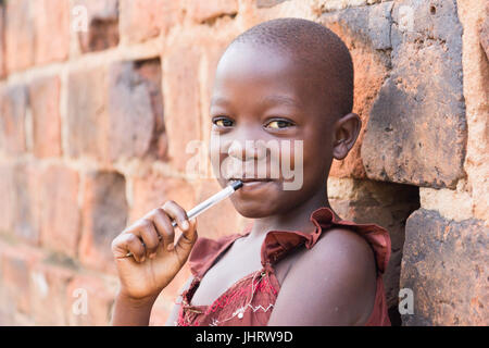 Un noir de 11 ans jeune fille ougandaise en souriant et tenant un stylo contre sa bouche et appuyé contre un mur de briques à l'appareil photo