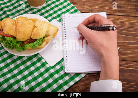 Croissant sandwich sur une soucoupe. Dans les mains d'un stylo écrit un plan d'activités à un ordinateur portable. Sur la table du salon. Déjeuner d'affaires concept. Banque D'Images