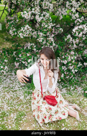 Jolie jeune femme à l'aide d'un téléphone mobile pour prendre des photos en extérieur. femme dans un chandail blanc et un jean bleu, assis sur l'herbe selfies et f Banque D'Images