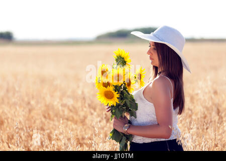 Portrait d'une belle jeune femme avec chapeau dans le champ de blé floue et le ciel en arrière-plan, qui tenait un bouquet de tournesols et enjo Banque D'Images