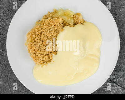 Crumble aux pommes dessert servi chaud avec de la crème anglaise contre une tuile ardoise cuisine noire Banque D'Images