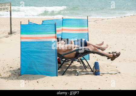 Boscombe, Bournemouth, Dorset, Royaume-Uni, 18th juillet 2017. Couple allongé sur la plage sur des chaises inclinables derrière le pare-brise. Banque D'Images
