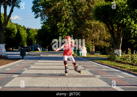 Jolie petite fille en tissu rouge à l'apprentissage à l'extérieur du patin sur belle journée d'été dans le parc de la ville. Banque D'Images