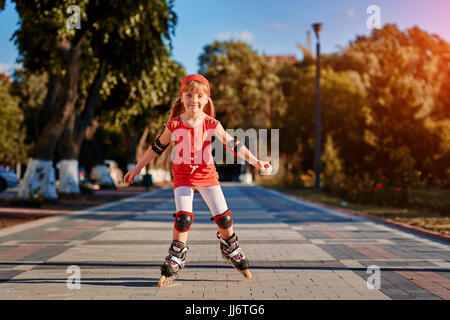 Jolie petite fille en tissu rouge à l'apprentissage à l'extérieur du patin sur belle journée d'été dans le parc de la ville. Banque D'Images