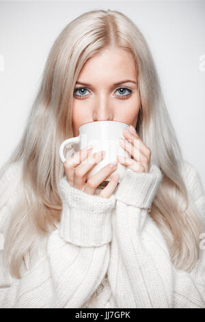 Jeune femme blonde vêtue de blanc grand pull en cachemire sur fond gris, de boire un thé de la pac Banque D'Images