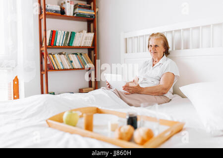 Happy elderly woman having breakfast in bed, tout en utilisant son tablet Banque D'Images