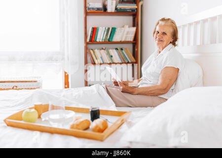 Happy elderly woman having breakfast in bed, tout en utilisant son tablet Banque D'Images