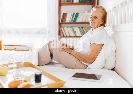 Happy elderly woman having breakfast in bed, tout en utilisant son tablet Banque D'Images