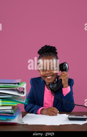 Portrait of smiling businesswoman talking on telephone tout en faisant des formalités administratives de bureau contre pink wall in office Banque D'Images