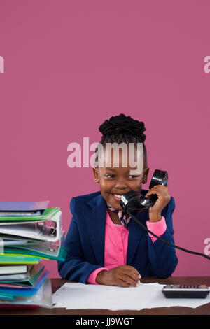 Smiling businesswoman talking on telephone tout en faisant des formalités administratives de bureau contre pink wall in office Banque D'Images