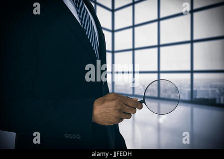 Mid section of businessman holding magnifying glass contre chambre avec grande fenêtre montrant city Banque D'Images